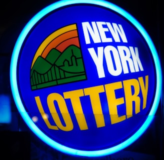 A bright, circular neon sign with a blue background, featuring the words 'New York Lottery' in bold white and yellow letters. The sign includes a colorful illustration of a landscape with green mountains, an orange-red sunset, and a bridge, all encapsulated within the circle.