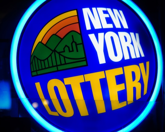 A bright, circular neon sign with a blue background, featuring the words 'New York Lottery' in bold white and yellow letters. The sign includes a colorful illustration of a landscape with green mountains, an orange-red sunset, and a bridge, all encapsulated within the circle.