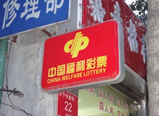 A red sign displaying the text 'China Welfare Lottery' in both Chinese and English, mounted on the exterior of a building. The surroundings include a blue sign with Chinese characters and a partial tree trunk visible on the right side.