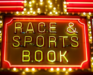 Bright neon lights on a marquee sign display the words 'RACE & SPORTS BOOK'. The sign is surrounded by rows of yellow and white lights against a red background, evoking a vibrant, energetic atmosphere typically associated with casinos or entertainment venues.