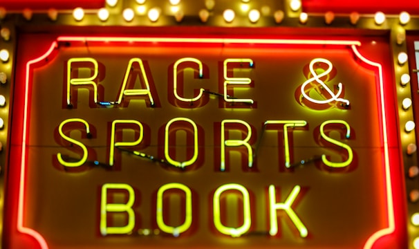 Bright neon lights on a marquee sign display the words 'RACE & SPORTS BOOK'. The sign is surrounded by rows of yellow and white lights against a red background, evoking a vibrant, energetic atmosphere typically associated with casinos or entertainment venues.