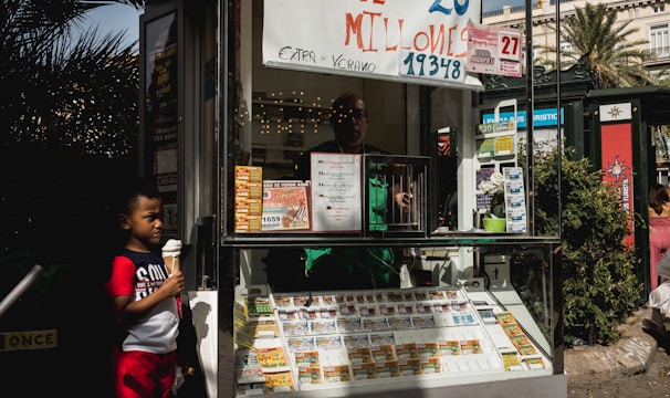 A child holding an ice cream cone stands outside a small kiosk or lottery ticket booth. The booth displays various colorful lottery tickets in the window. There are signs advertising a significant lottery prize amount in Spanish. A person can be seen partially obscured inside the booth, and plants are visible around the area, suggesting an outdoor setting.
