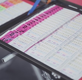 A collection of lottery tickets is organized neatly in a metal tray. The tickets display various numbers and have color-coded sections, primarily in pink and white. Some tickets have vibrant green sections, adding contrast to the overall layout.