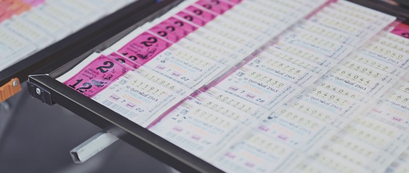 A collection of lottery tickets is organized neatly in a metal tray. The tickets display various numbers and have color-coded sections, primarily in pink and white. Some tickets have vibrant green sections, adding contrast to the overall layout.