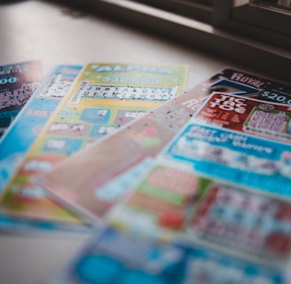 A selection of colorful lottery scratch-off tickets placed on a flat surface near a window. The image has a shallow depth of field, focusing on the tickets while blurring the background. The tickets display various games and prize amounts.