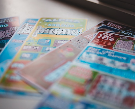 A selection of colorful lottery scratch-off tickets placed on a flat surface near a window. The image has a shallow depth of field, focusing on the tickets while blurring the background. The tickets display various games and prize amounts.