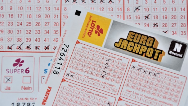 Three lottery tickets are displayed, including Eurojackpot and Super 6. Various numbers are marked on the tickets, with some crossed out and others checked. The Eurojackpot ticket is partially visible with the logo prominently displayed, and a ticket number is visible on one of the tickets.