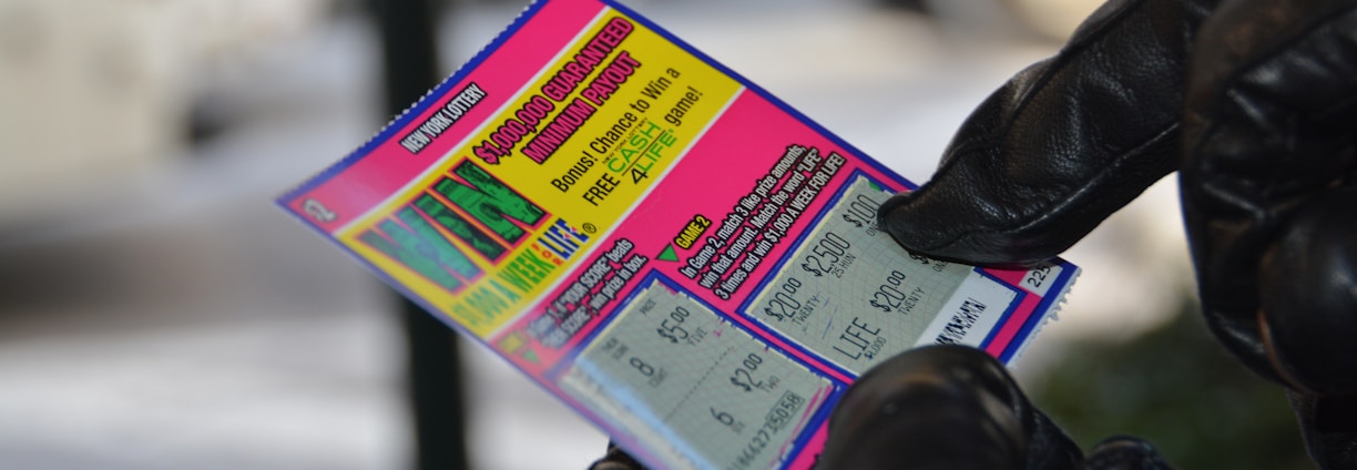 A person wearing black gloves is holding a colorful lottery scratch-off ticket. The ticket includes information about a $1,000,000 guaranteed minimum payout and features multiple games with various prize amounts.