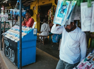 A street vendor stall displaying an array of lottery tickets clipped to a bright blue kiosk. A man in a red shirt leans on the counter, appearing thoughtful, while another man in a white shirt adjusts his hat and walks by. A variety of products hangs from strings above, and a colorful display fills the background with vibrant signs and packets. The scene suggests a busy, lively marketplace.