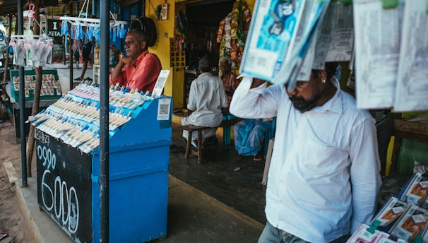 A street vendor stall displaying an array of lottery tickets clipped to a bright blue kiosk. A man in a red shirt leans on the counter, appearing thoughtful, while another man in a white shirt adjusts his hat and walks by. A variety of products hangs from strings above, and a colorful display fills the background with vibrant signs and packets. The scene suggests a busy, lively marketplace.
